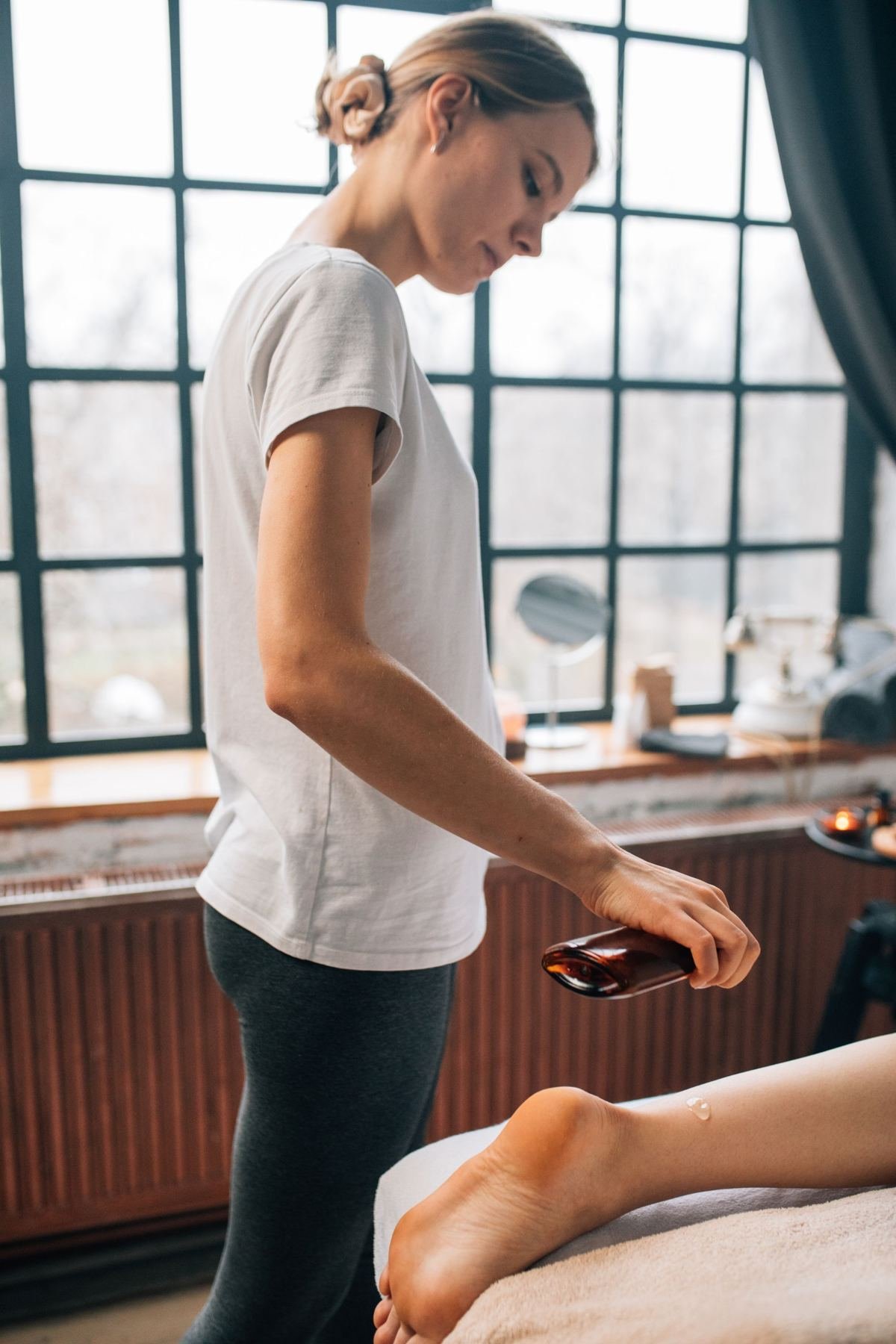 Une praticienne verse de l'huile de massage dans sa main, près d'une cliente allongée dans une pièce lumineuse